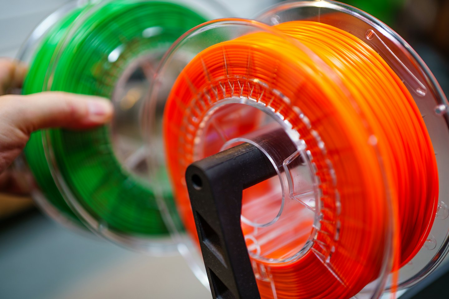 Hand holds a spool of green 3D printer filament next to an orange spool.